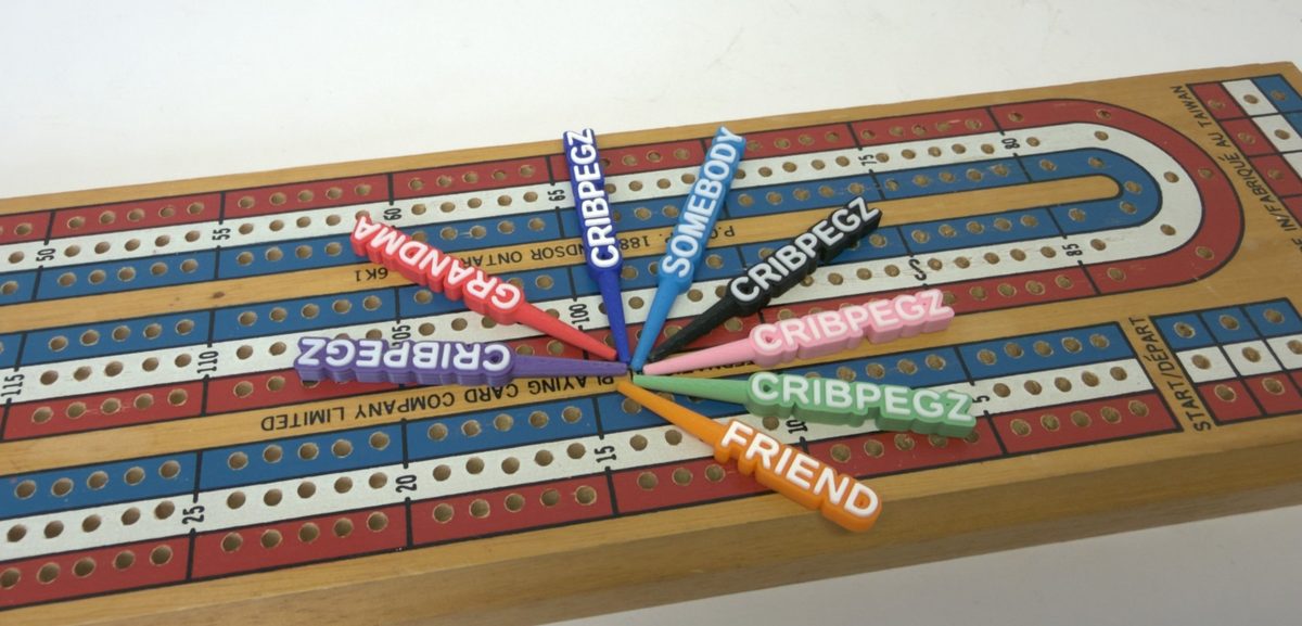 Custom cribbage pegs fanned out on a cribbage board: CRIBPEGZ in purple, dark blue, black, pink and green, GRANDMA in red, SOMEBODY in light blue and FRIEND in orange