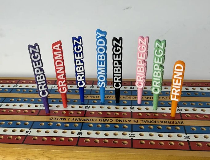 Eight custom cribbage pegs standing in a board: CRIBPEGZ in purple, dark blue, black, pink and green, GRANDMA in red, SOMEBODY in light blue, FRIEND in orange